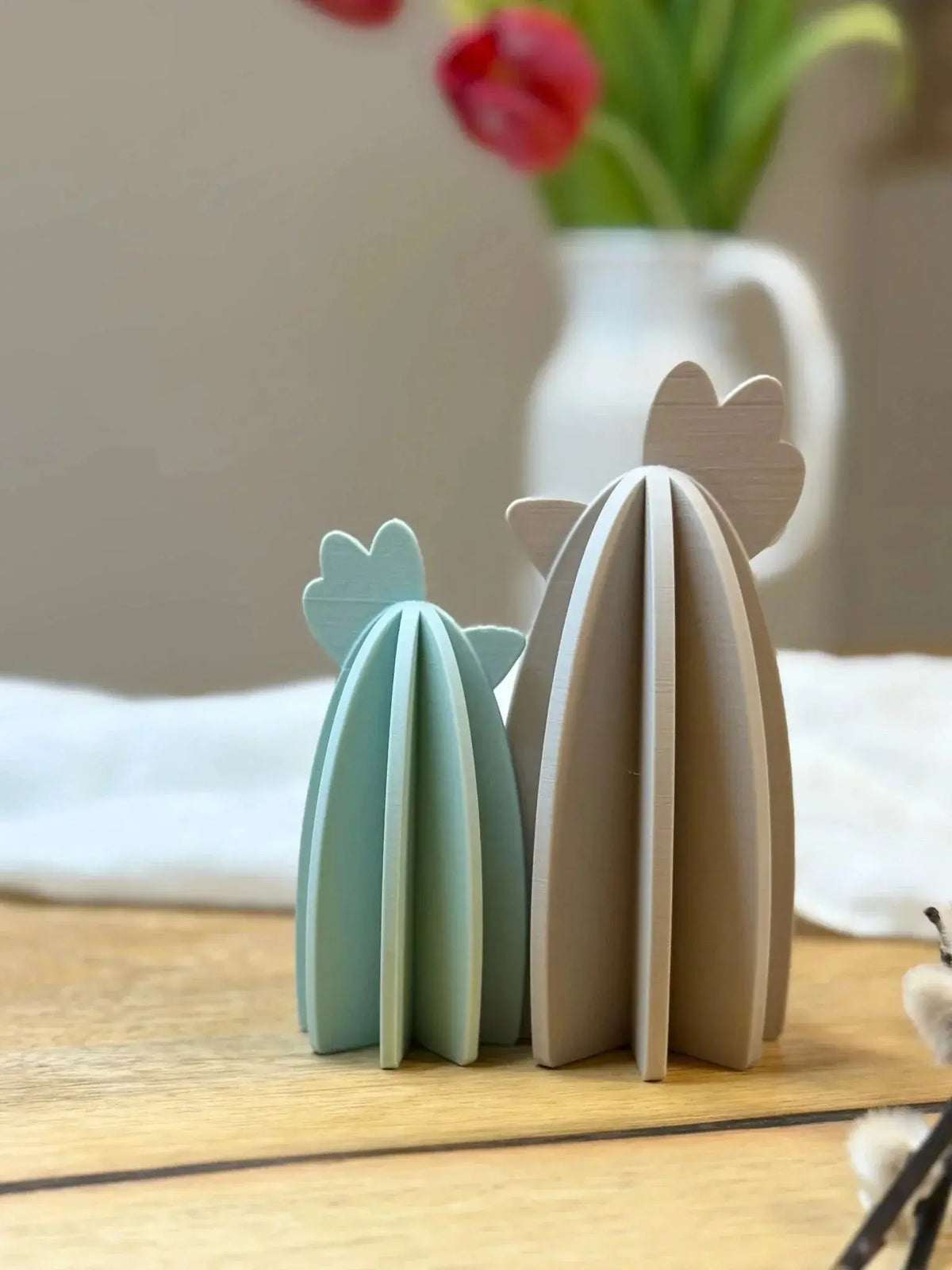 Zwei dekorative, pastellfarbene Holz-Kakteen auf Tisch, Vase mit Tulpen im Hintergrund.