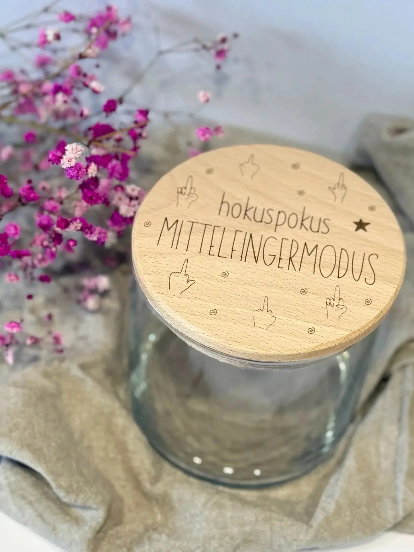 Personalisierbares Vorratsglas mit graviertem Holzdeckel 'hokuspokus Mittelfingermodus', daneben pinke Trockenblumen.